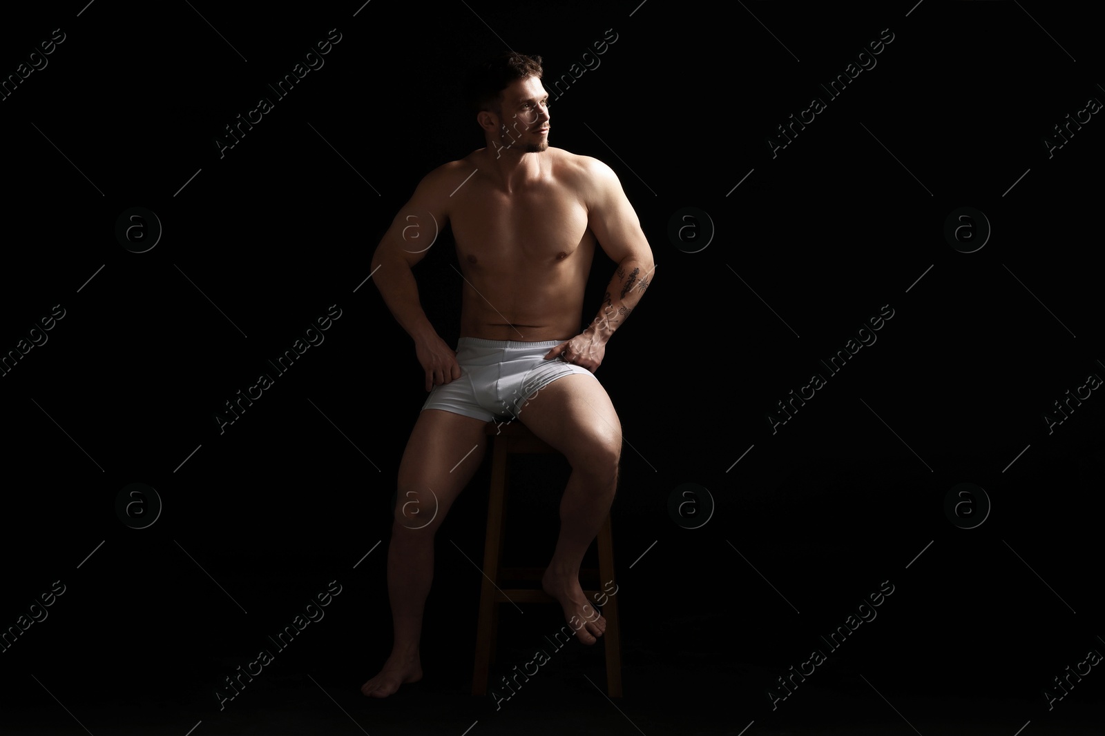Young man in stylish white underwear on chair against black background Photo of Young man in stylish white underwear on chair against black background