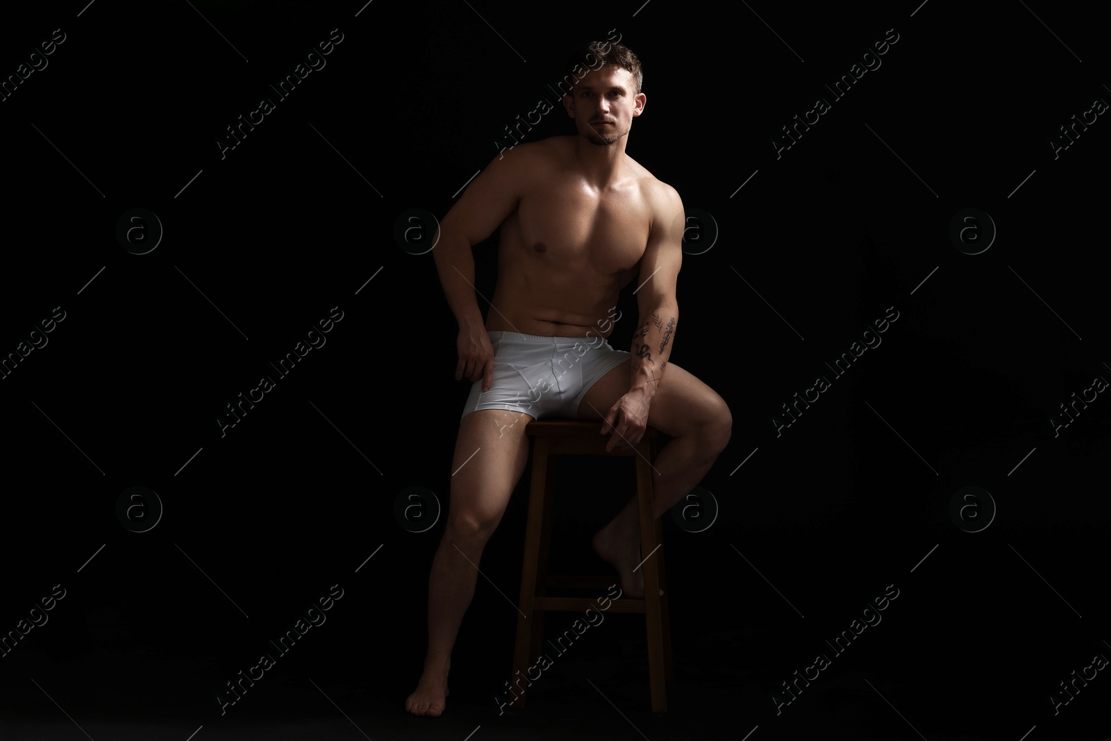 Young man in stylish white underwear on chair against black background Photo of Young man in stylish white underwear on chair against black background