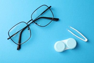 Photo of Contact lens case, tweezers and glasses on light blue background, flat lay