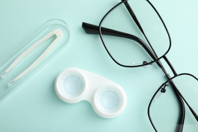 Photo of Contact lenses in case, tweezers and glasses on light turquoise background, flat lay