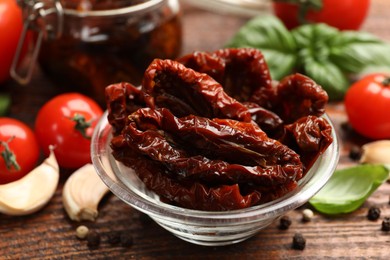 Photo of Delicious sun dried tomatoes in glass bowl, fresh vegetables and spices on wooden table, closeup