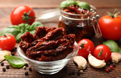 Photo of Delicious sun dried tomatoes in glass dishware, fresh vegetables and spices on wooden table, closeup