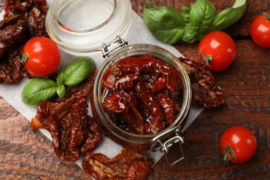 Photo of Delicious sun dried tomatoes in glass jar, fresh vegetables and basil on wooden table, flat lay
