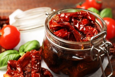 Photo of Delicious sun dried tomatoes in glass jar, fresh vegetables and basil on table, closeup