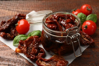 Photo of Delicious sun dried tomatoes in glass jar, fresh vegetables and basil on wooden table, closeup