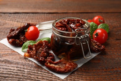 Photo of Delicious sun dried tomatoes in glass jar, fresh vegetables and basil on wooden table, closeup