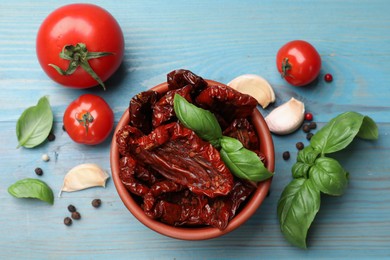 Photo of Delicious sun dried tomatoes in bowl, fresh vegetables and spices on light blue wooden table, flat lay