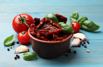 Photo of Delicious sun dried tomatoes in bowl, fresh vegetables and spices on light blue wooden table, closeup