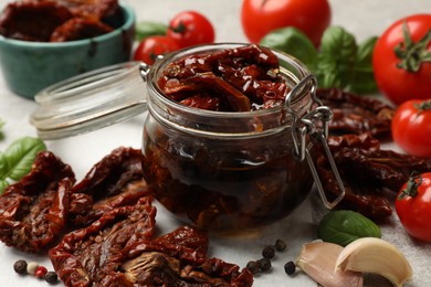 Photo of Delicious sun dried tomatoes in glass jar, fresh vegetables and spices on gray table, closeup