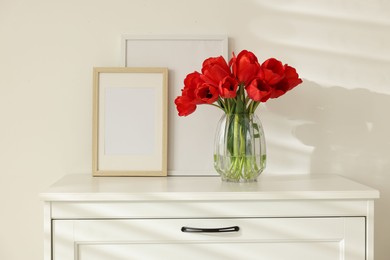 Photo of Beautiful red tulips in vase and frames on dresser indoors