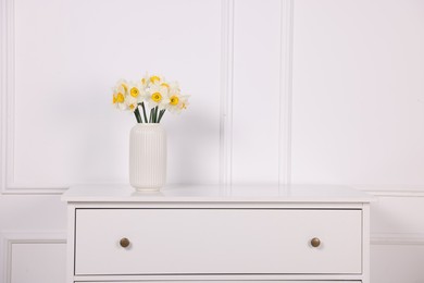 Photo of Beautiful daffodils in vase on dresser near white wall at home