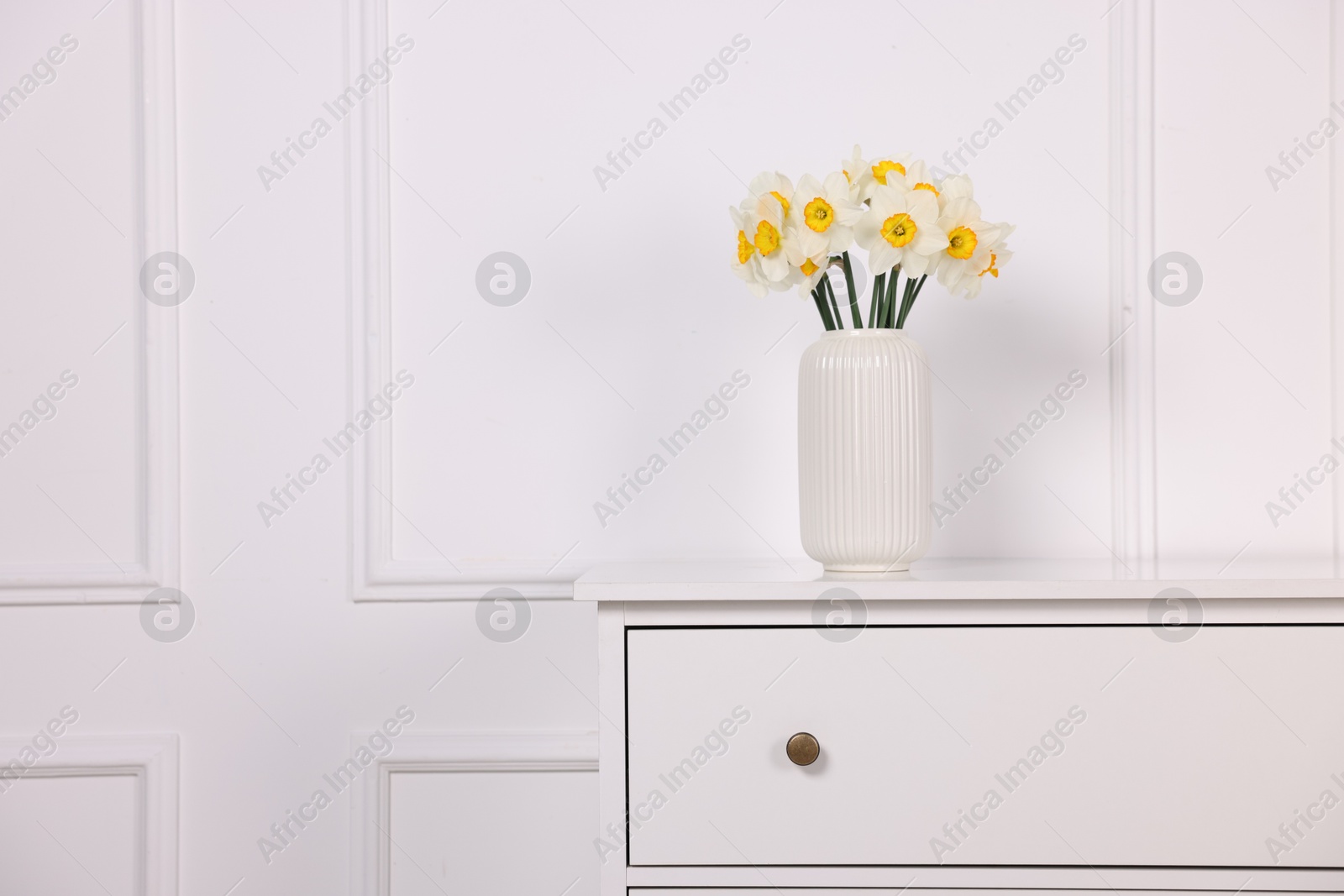Beautiful daffodils in vase on dresser near white wall at home, space for text Photo of Beautiful daffodils in vase on dresser near white wall at home, space for text