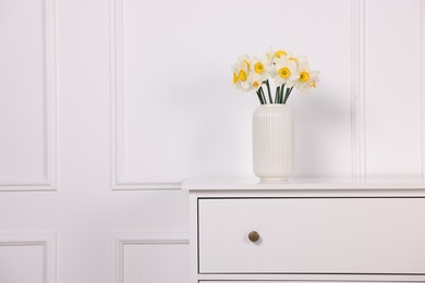 Photo of Beautiful daffodils in vase on dresser near white wall at home, space for text