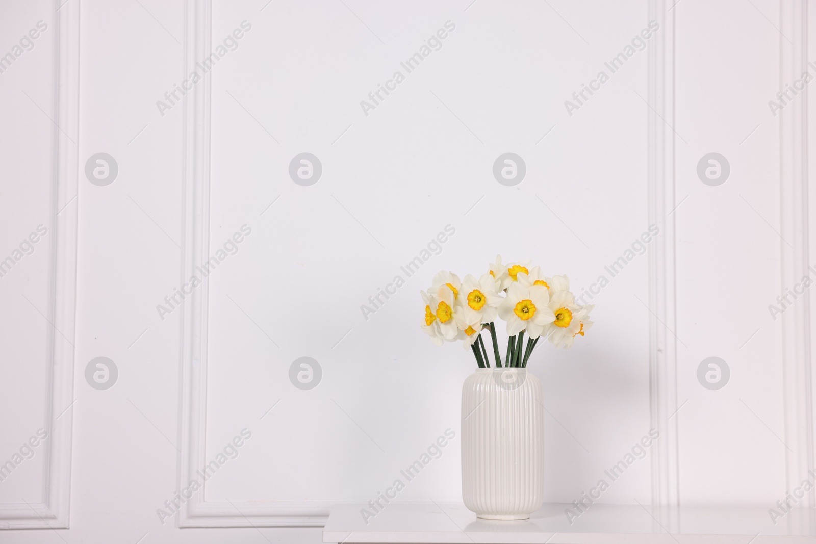 Beautiful daffodils in vase on table near white wall at home, space for text Photo of Beautiful daffodils in vase on table near white wall at home, space for text