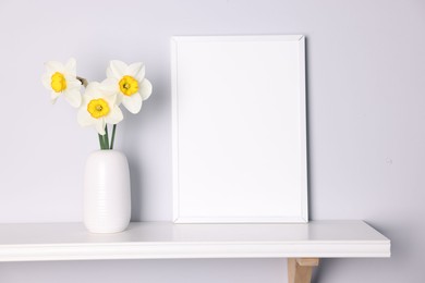 Photo of Beautiful daffodils in vase and picture frame on white shelf indoors