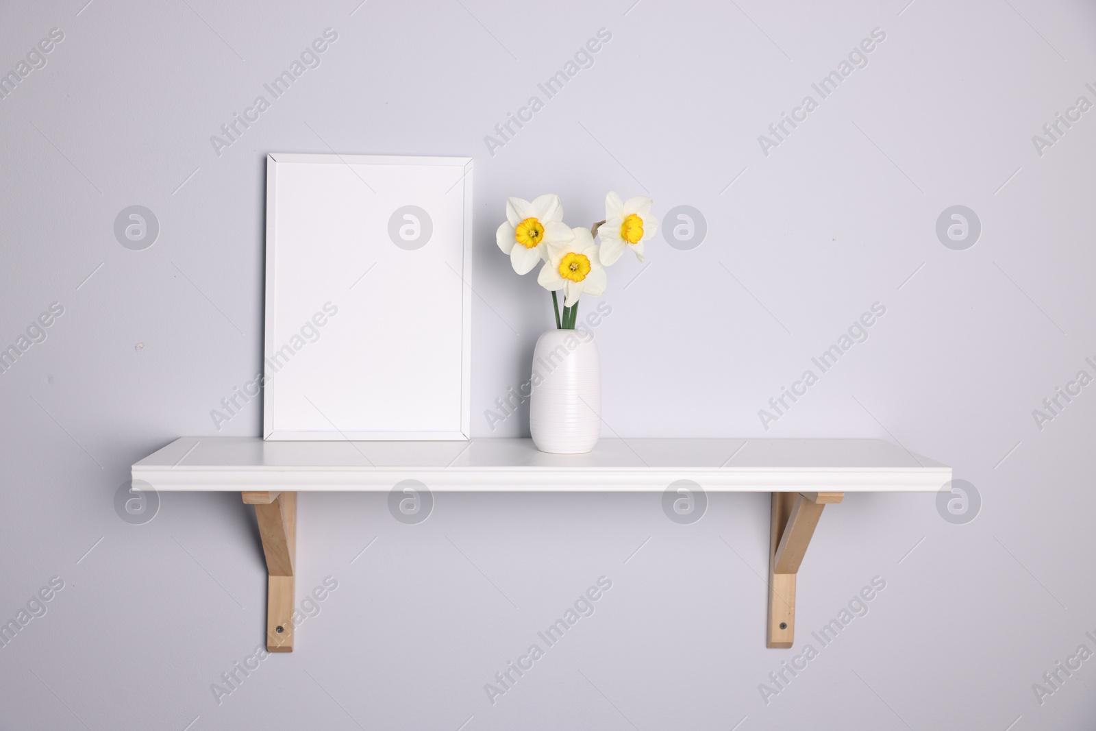 Beautiful daffodils in vase and picture frame on white shelf indoors Photo of Beautiful daffodils in vase and picture frame on white shelf indoors