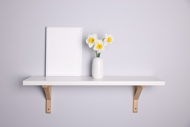 Photo of Beautiful daffodils in vase and picture frame on white shelf indoors