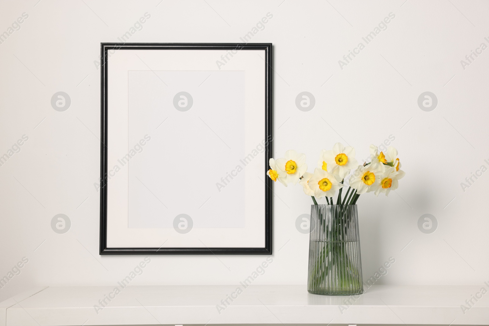 Beautiful daffodils in vase on white table and picture frame indoors Photo of Beautiful daffodils in vase on white table and picture frame indoors