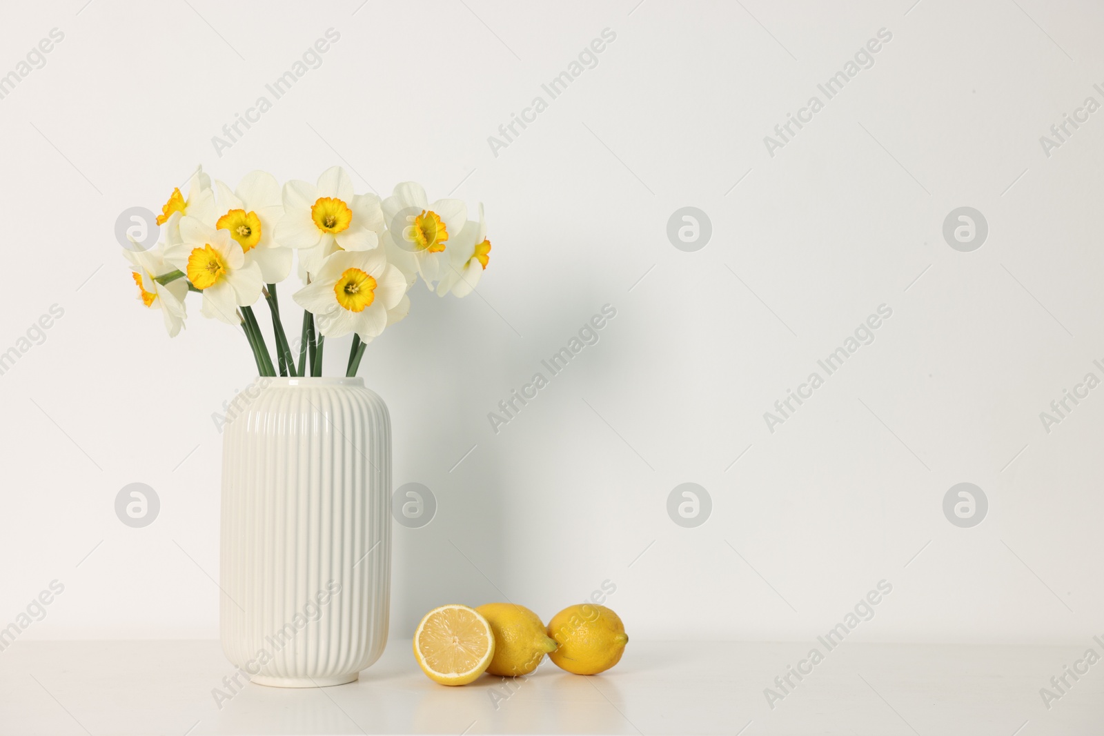 Beautiful daffodils in vase and lemons on white table indoors, space for text Photo of Beautiful daffodils in vase and lemons on white table indoors, space for text