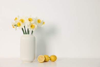 Photo of Beautiful daffodils in vase and lemons on white table indoors, space for text