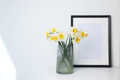 Photo of Beautiful daffodils in vase and picture frame on white wooden table indoors, space for text