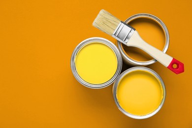 Image of Cans with paint in shades of yellow and brush on orange background, top view. Space for text