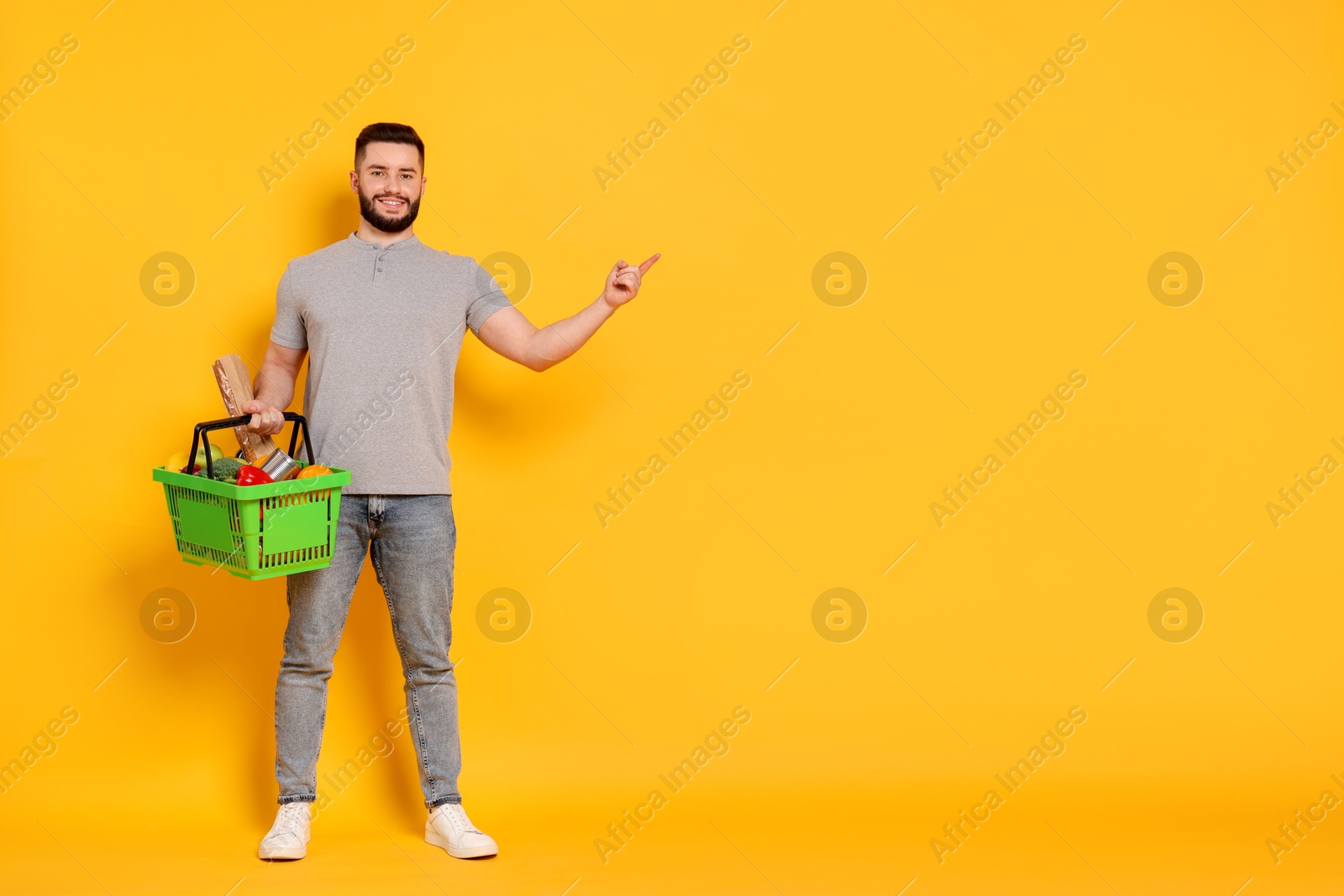Man with shopping basket full of products pointing at something on orange background, space for text Photo of Man with shopping basket full of products pointing at something on orange background, space for text