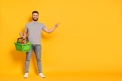 Photo of Man with shopping basket full of products pointing at something on orange background, space for text