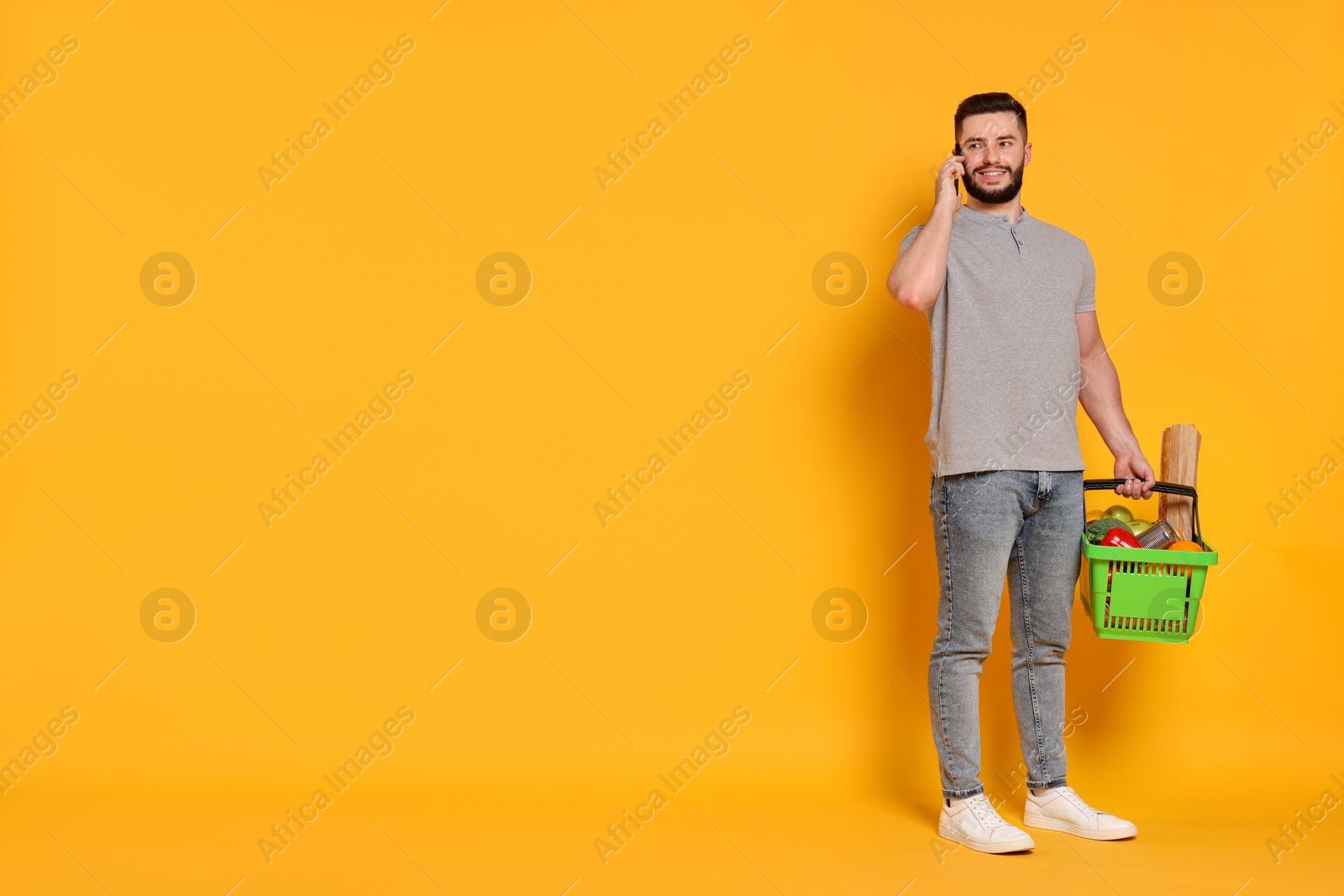 Man with shopping basket full of products talking on smartphone against orange background, space for text Photo of Man with shopping basket full of products talking on smartphone against orange background, space for text