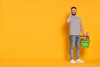 Photo of Man with shopping basket full of products talking on smartphone against orange background, space for text