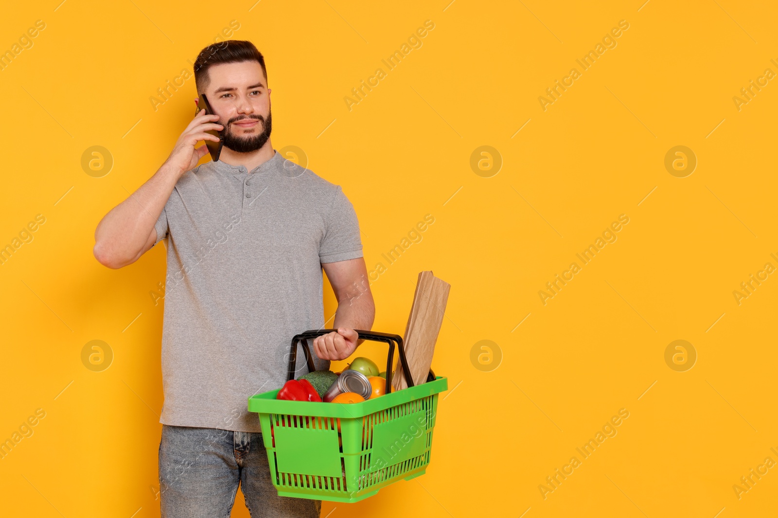 Man with shopping basket full of products talking on smartphone against orange background, space for text Photo of Man with shopping basket full of products talking on smartphone against orange background, space for text