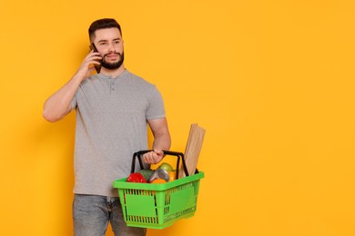 Photo of Man with shopping basket full of products talking on smartphone against orange background, space for text