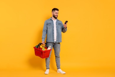 Photo of Man with shopping basket full of products and smartphone on orange background