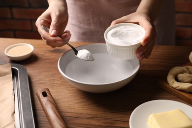 Photo of Making pretzels. Woman adding baking soda into bowl of water at wooden table, closeup