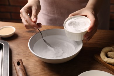 Photo of Making pretzels. Woman adding baking soda into bowl of water at wooden table, closeup