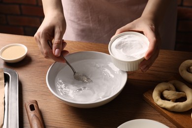 Photo of Making pretzels. Woman adding baking soda into bowl of water at wooden table, closeup
