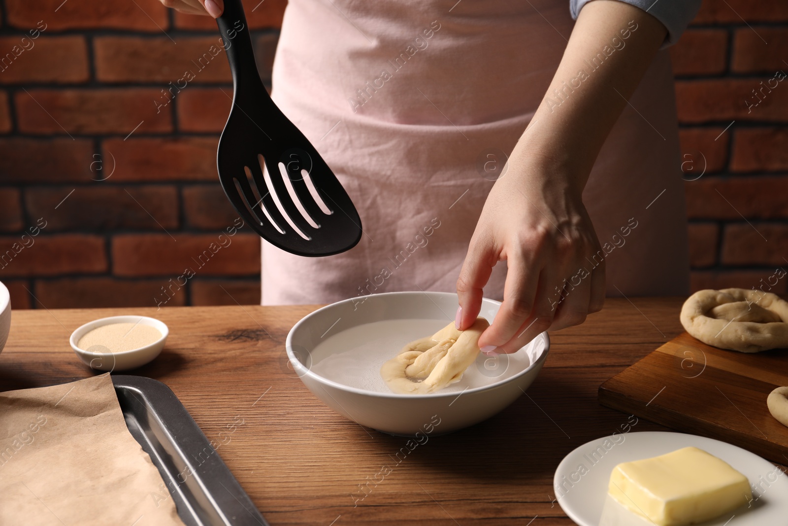 Woman putting uncooked pretzel into bowl of water at wooden table, closeup Photo of Woman putting uncooked pretzel into bowl of water at wooden table, closeup