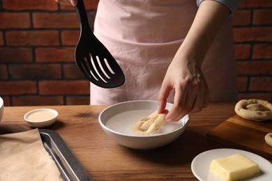 Photo of Woman putting uncooked pretzel into bowl of water at wooden table, closeup