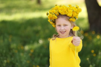 Photo of Cute little girl with beautiful flower wearing wreath of dandelions in park, space for text