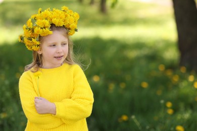 Photo of Cute little girl with beautiful flower wearing wreath of dandelions in park, space for text