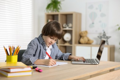 Photo of Cute little boy doing homework with laptop at table indoors