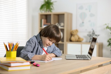 Photo of Cute little boy doing homework with laptop at table indoors