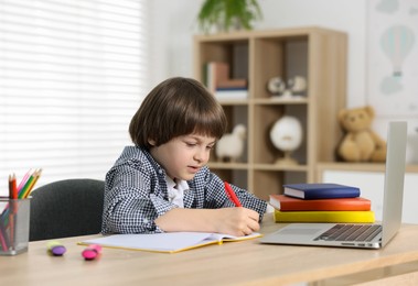 Photo of Cute little boy doing homework with laptop at table indoors