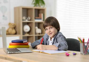 Photo of Cute little boy doing homework with laptop at table indoors