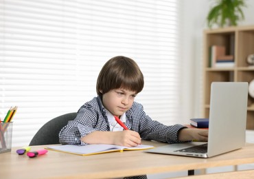 Photo of Cute little boy doing homework with laptop at table indoors