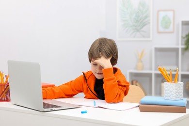 Photo of Tired little boy doing homework with laptop at desk indoors