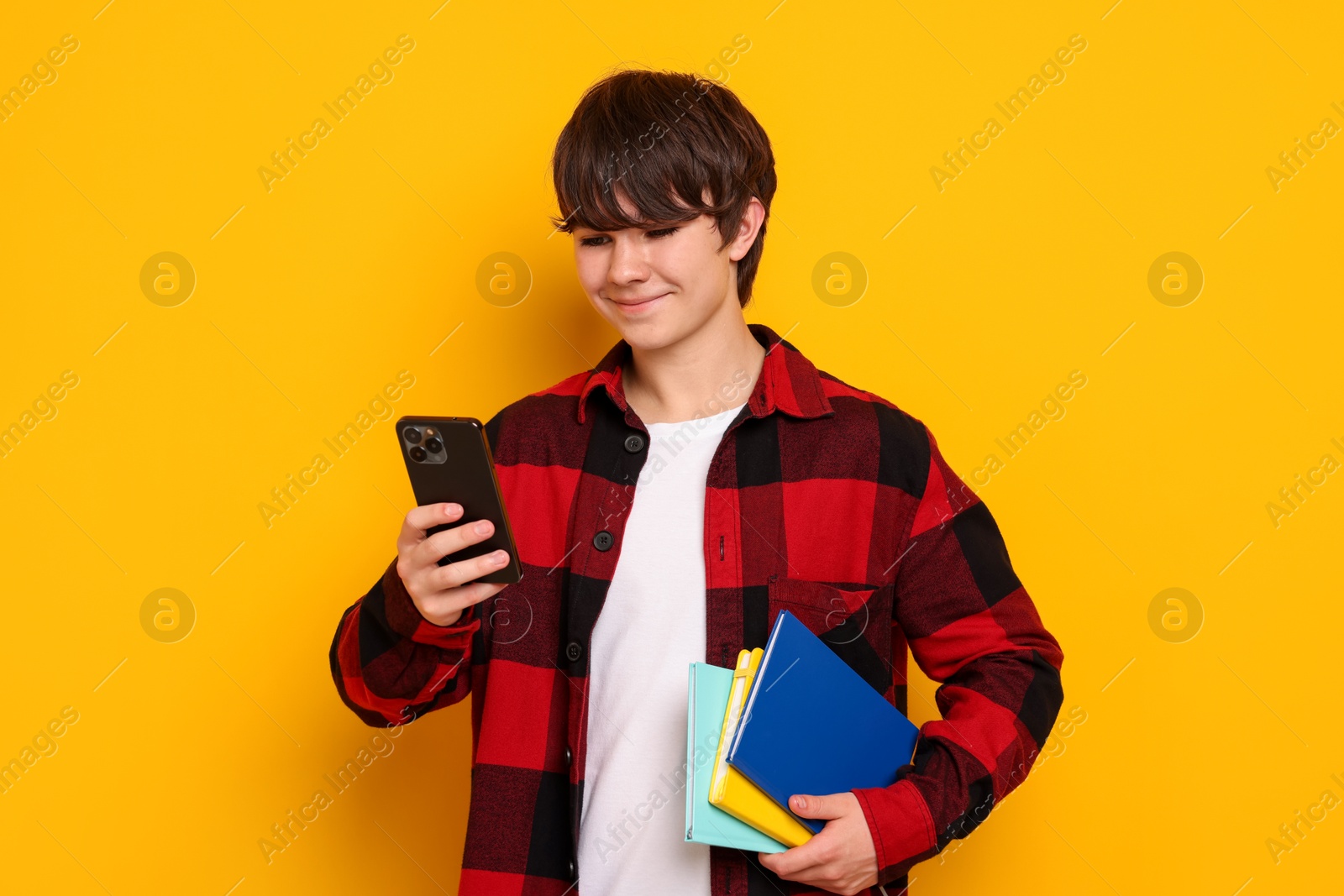 Teenage boy with smartphone and notebooks on orange background Photo of Teenage boy with smartphone and notebooks on orange background