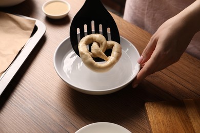 Photo of Woman putting uncooked pretzel into bowl of water at wooden table, closeup