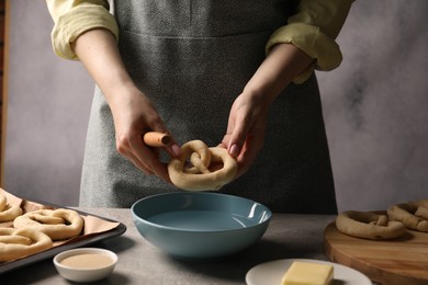 Photo of Woman putting uncooked pretzel into bowl of water at grey table, closeup