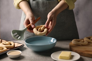 Photo of Woman putting uncooked pretzel into bowl of water at grey table, closeup